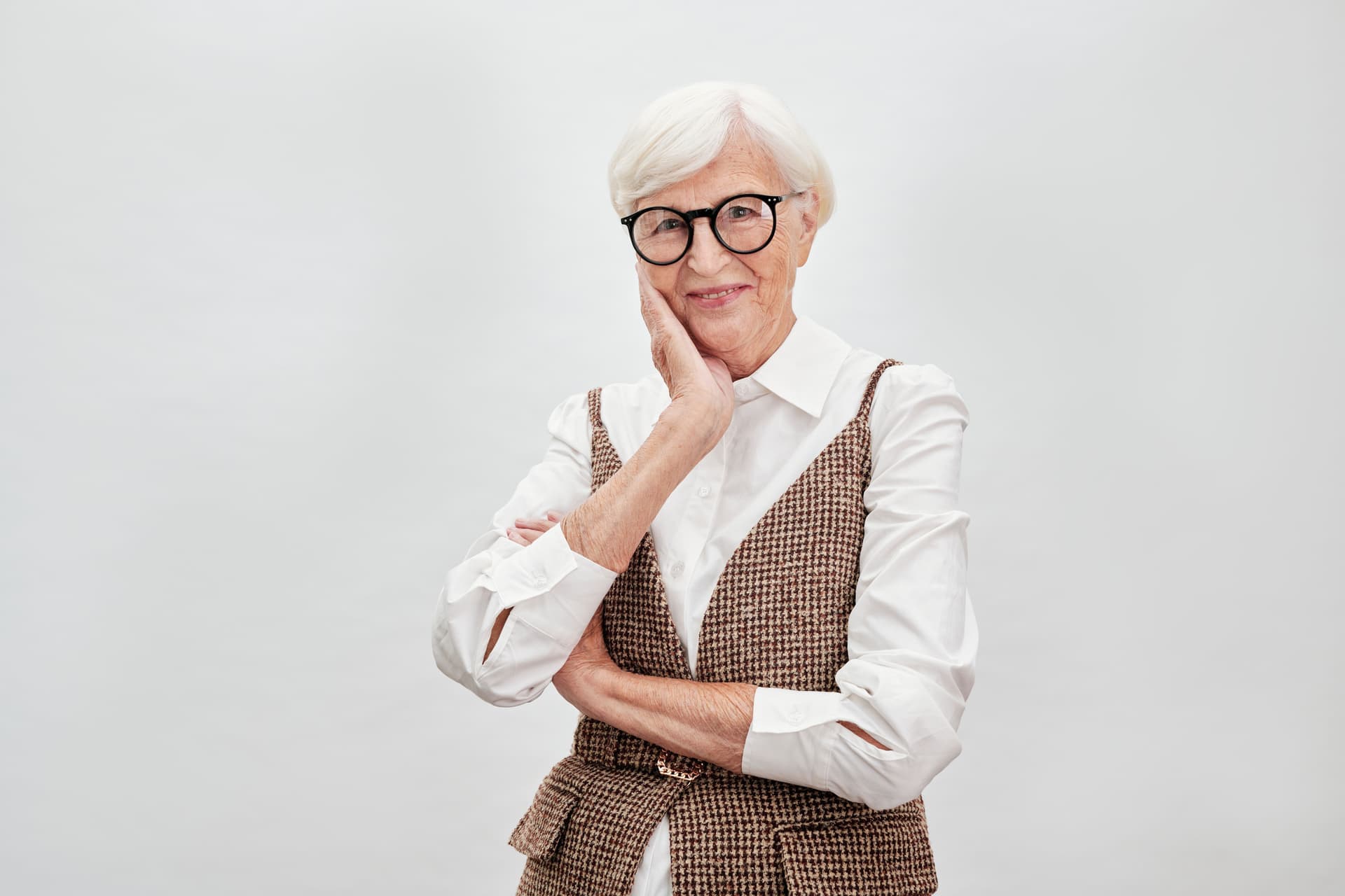 A smiling woman in her seventies with white hair and round glasses, photographed against a plain pale studio background