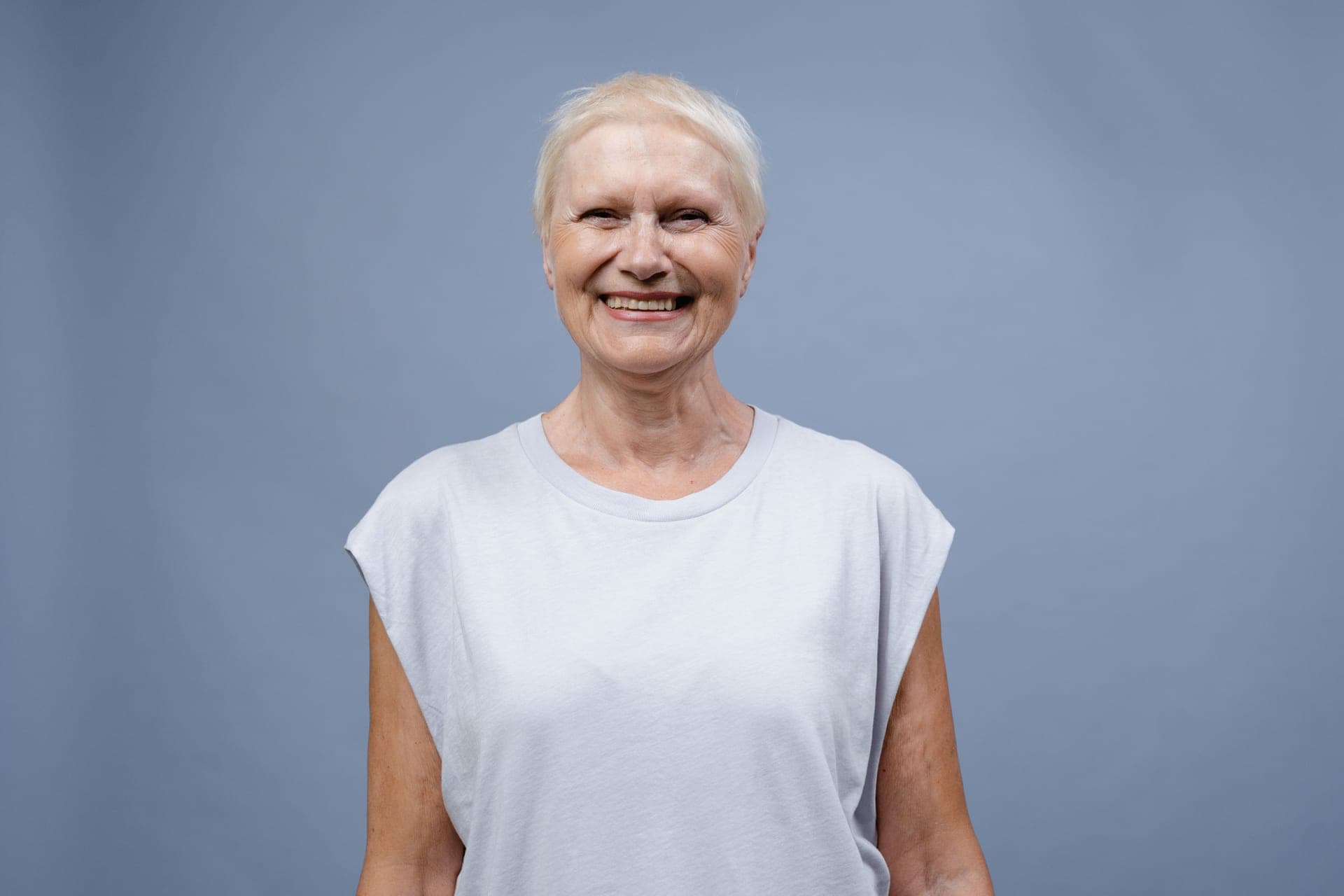 A smiling senior woman with short white hair, photographed against a plain studio backdrop